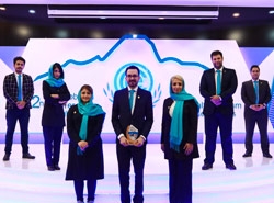 تیم آریامدتور با پنج مرد و دو زن با لباس مشکی و آبی در محل برگزاری دومین کنفرانس نمایشگاه رویداد اکو در حوزهی گردشگری سلامت یا مدیکال توریسم ایستادهاند و هادی شجاری در مقابل دیده میشود. پسزمینه صفحهای از ترکیب رنگهای سفید و بنفش است.
