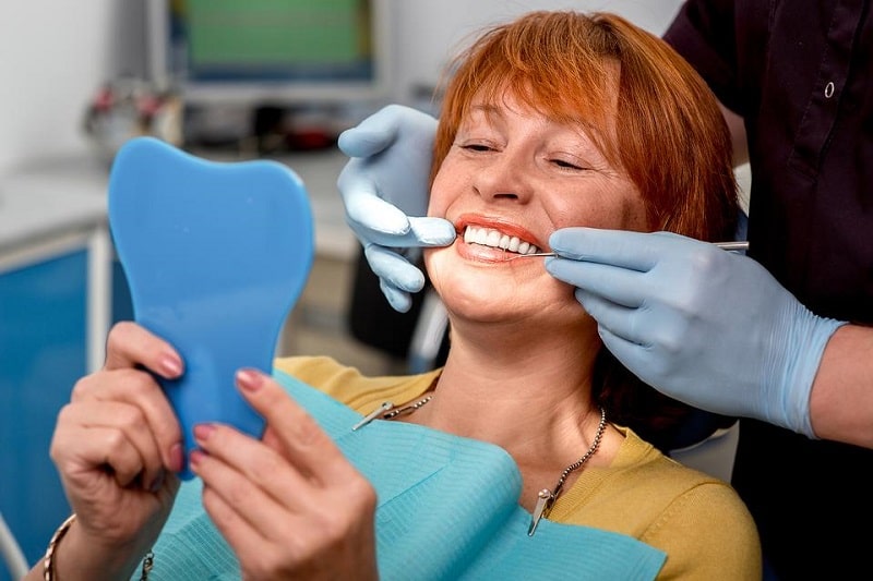 woman sitting in a dentist's chair looking in a mirror while her dentures are being checked by the dentist