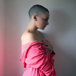 women in pink dress with shaved head putting her left hand on her chest indicating breast cancer