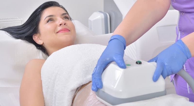 smiling woman lying on bed with a doctor placing a coolsculpting device on her belly