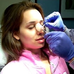 woman wearing pink jacket sitting in medical chair getting her nose bandage removed by a plastic surgeon who is wearing blue gloves