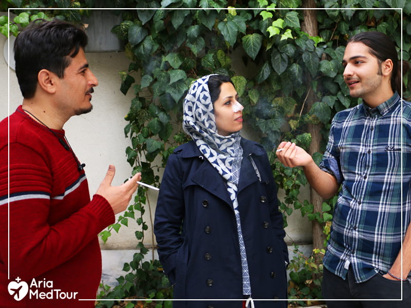 Two Iranian men smoking cigarettes and talking with a Muslim woman with a splint on her nose in a green space